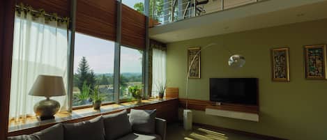 A modern living room with a sofa, a television mounted on the wall, and large windows offering a view of the outdoors.