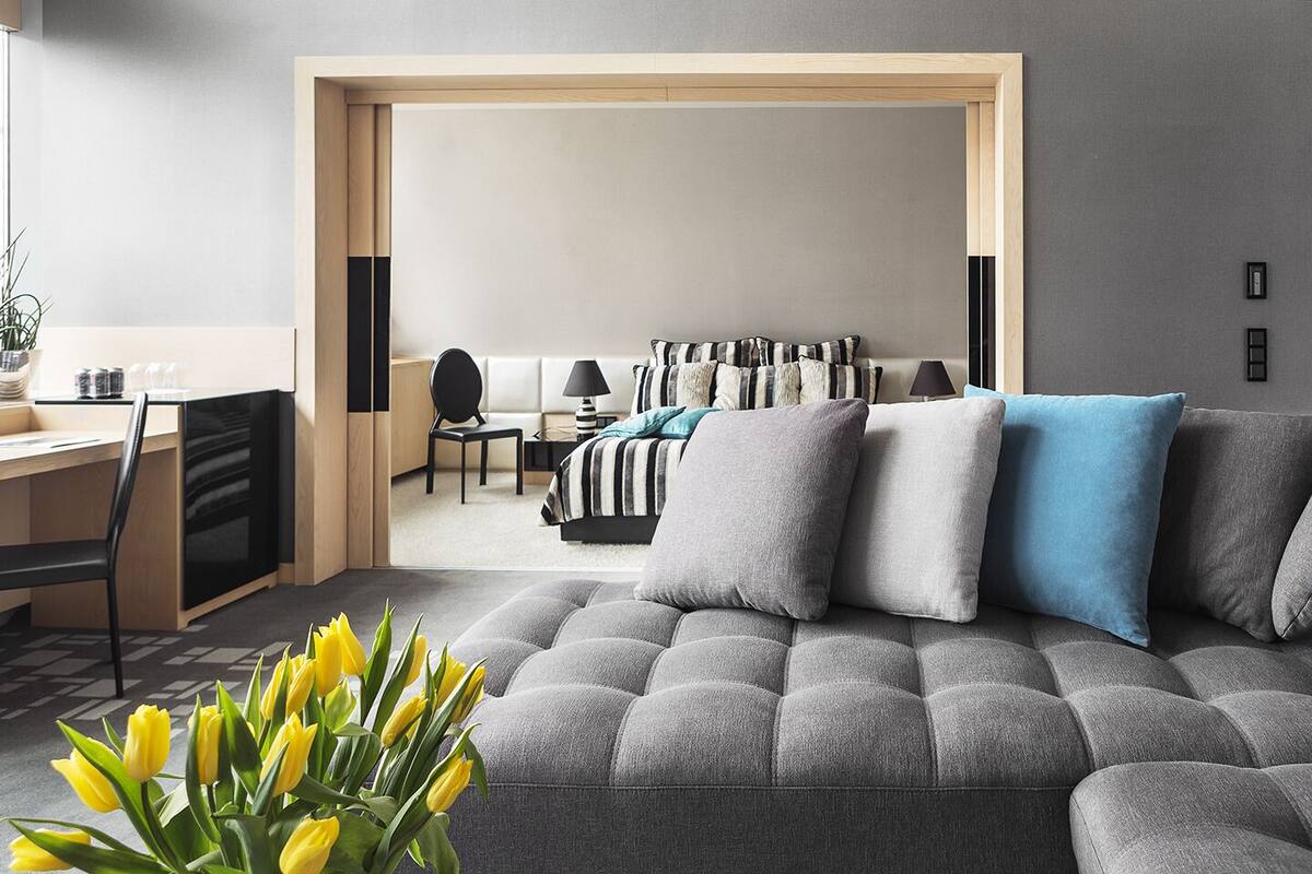 Una sala moderna con un sofá gris, un comedor con una silla negra y un dormitorio con colcha a rayas.