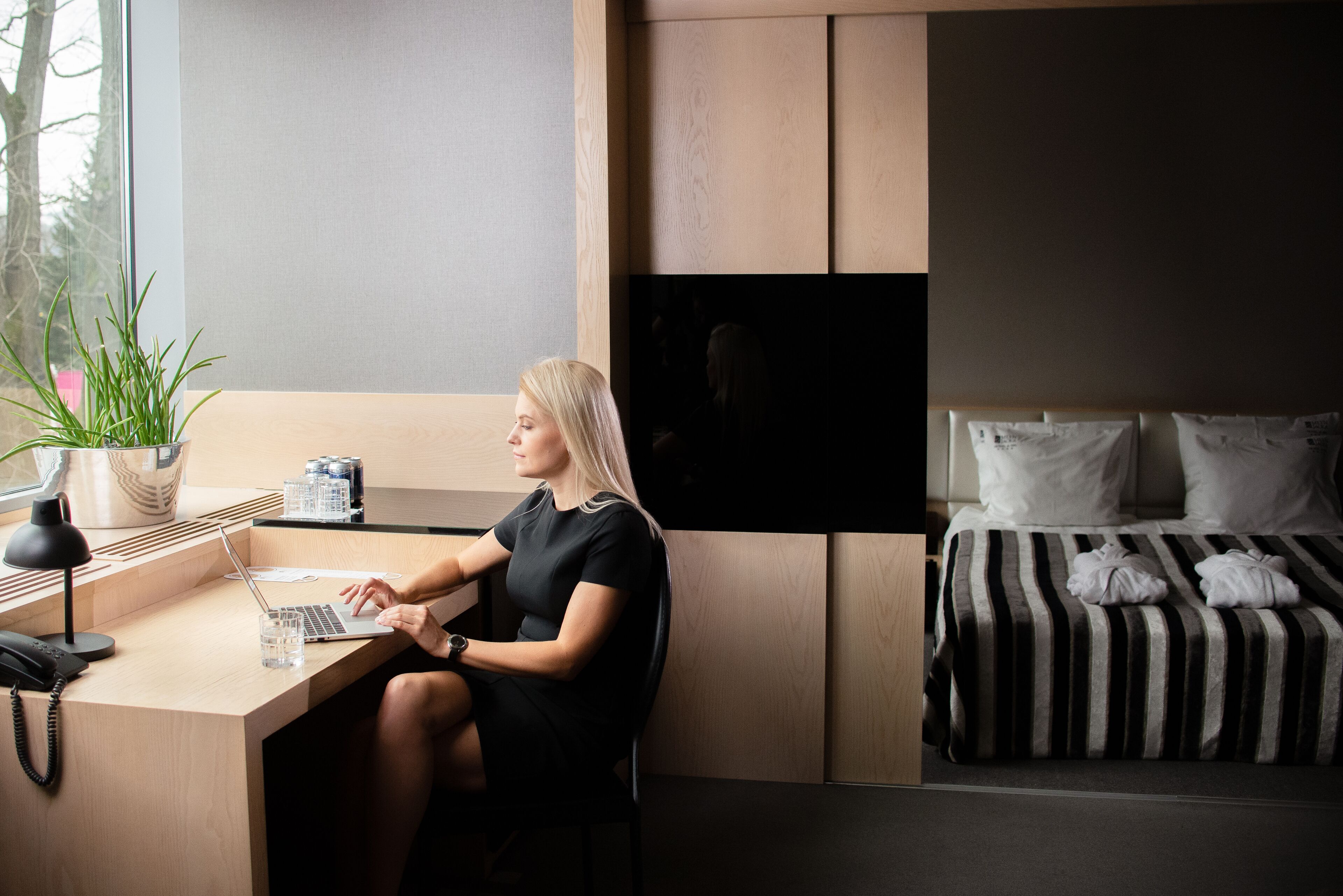 Habitación de hotel moderna con un escritorio de madera, una computadora portátil, una planta en maceta y una cama con ropa de cama a rayas.