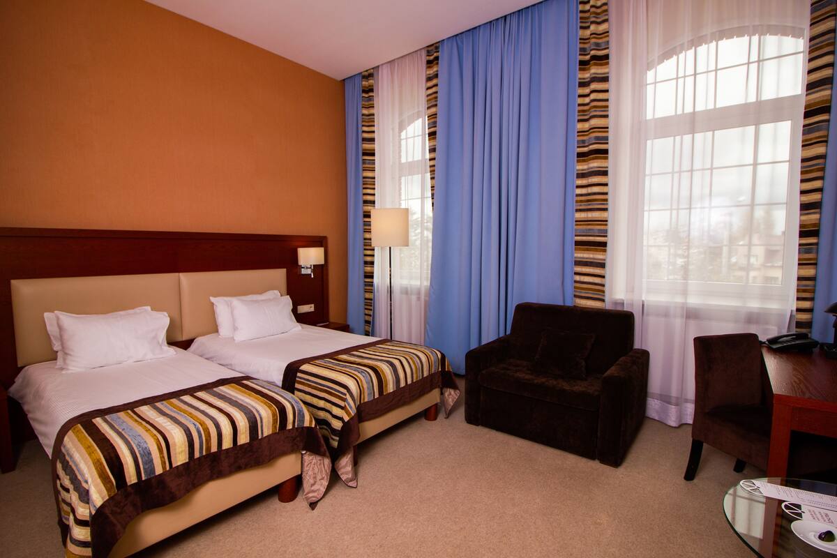 Una habitación de hotel con dos camas, un sillón marrón, un escritorio con lámpara y una ventana con cortinas.