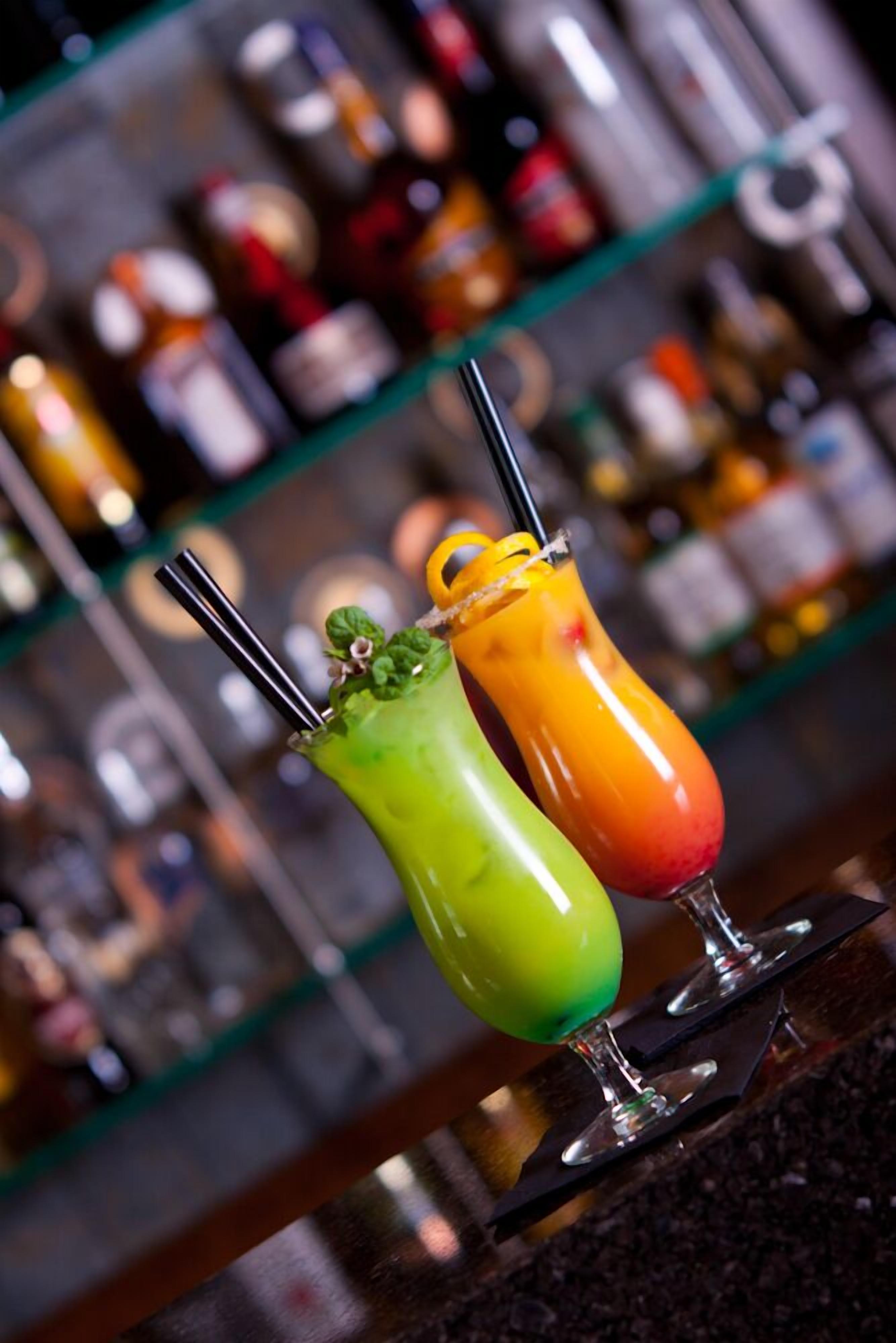 Deux cocktails colorés sur le comptoir d’un bar, avec un arrière-plan flou composé de différentes bouteilles.