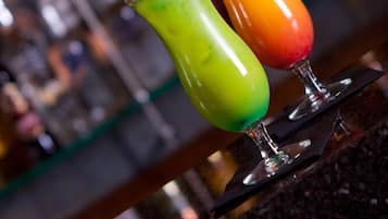 Deux cocktails colorés sur le comptoir d’un bar, avec un arrière-plan flou composé de différentes bouteilles.