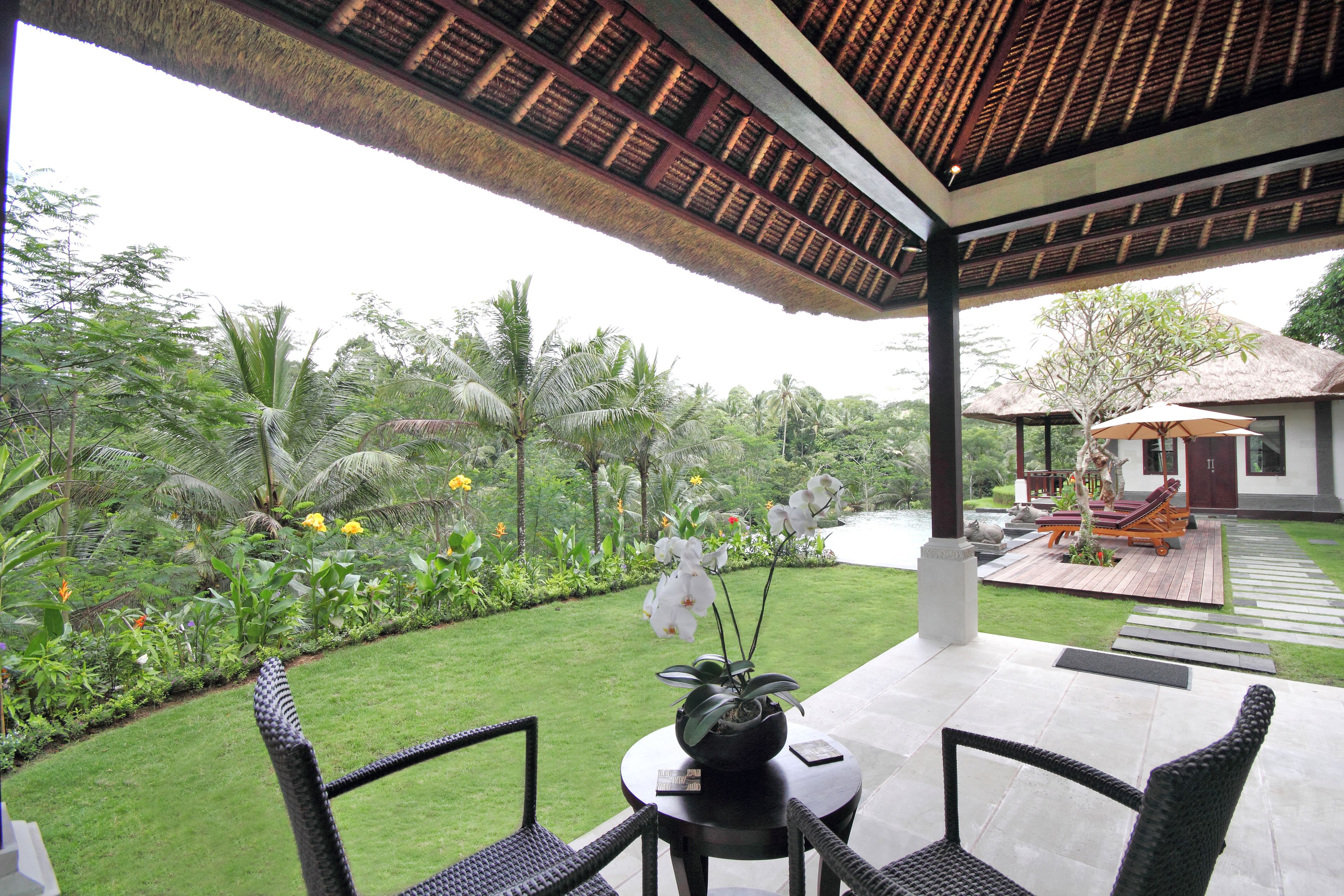 A covered outdoor area with a table and chairs, surrounded by lush greenery and a pathway leading to a building.