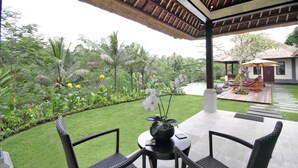 A covered outdoor area with a table and chairs, surrounded by lush greenery and a pathway leading to a building.