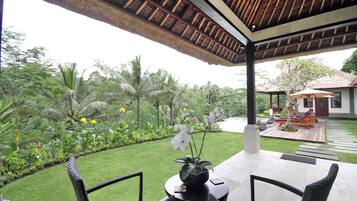 A covered outdoor area with a table and chairs, surrounded by lush greenery and a pathway leading to a building.