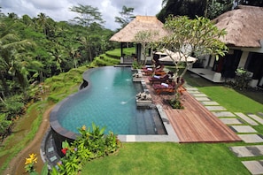 A swimming pool with a wooden deck, lounge chairs, and a thatched-roof structure. - Villa Amrita (Tegallalang)