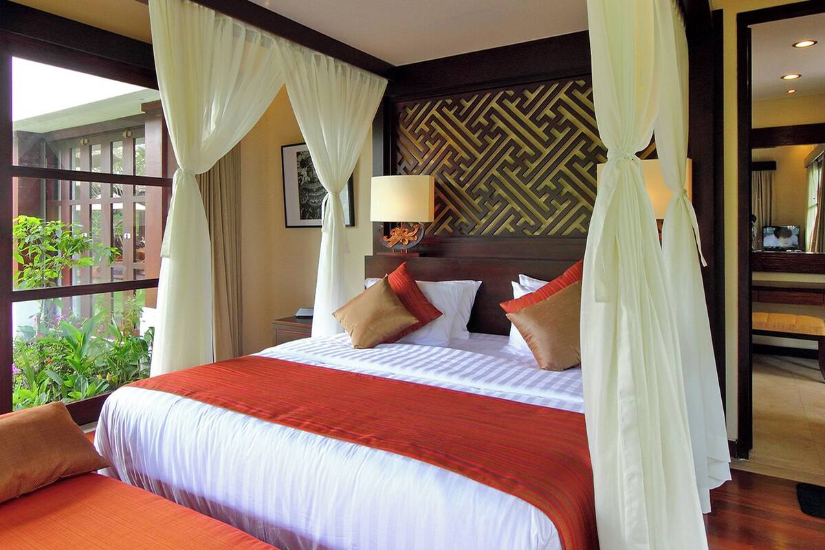 A hotel room with a large bed, a sitting area with a sofa, a bathroom with a mirror, and a view of a garden.