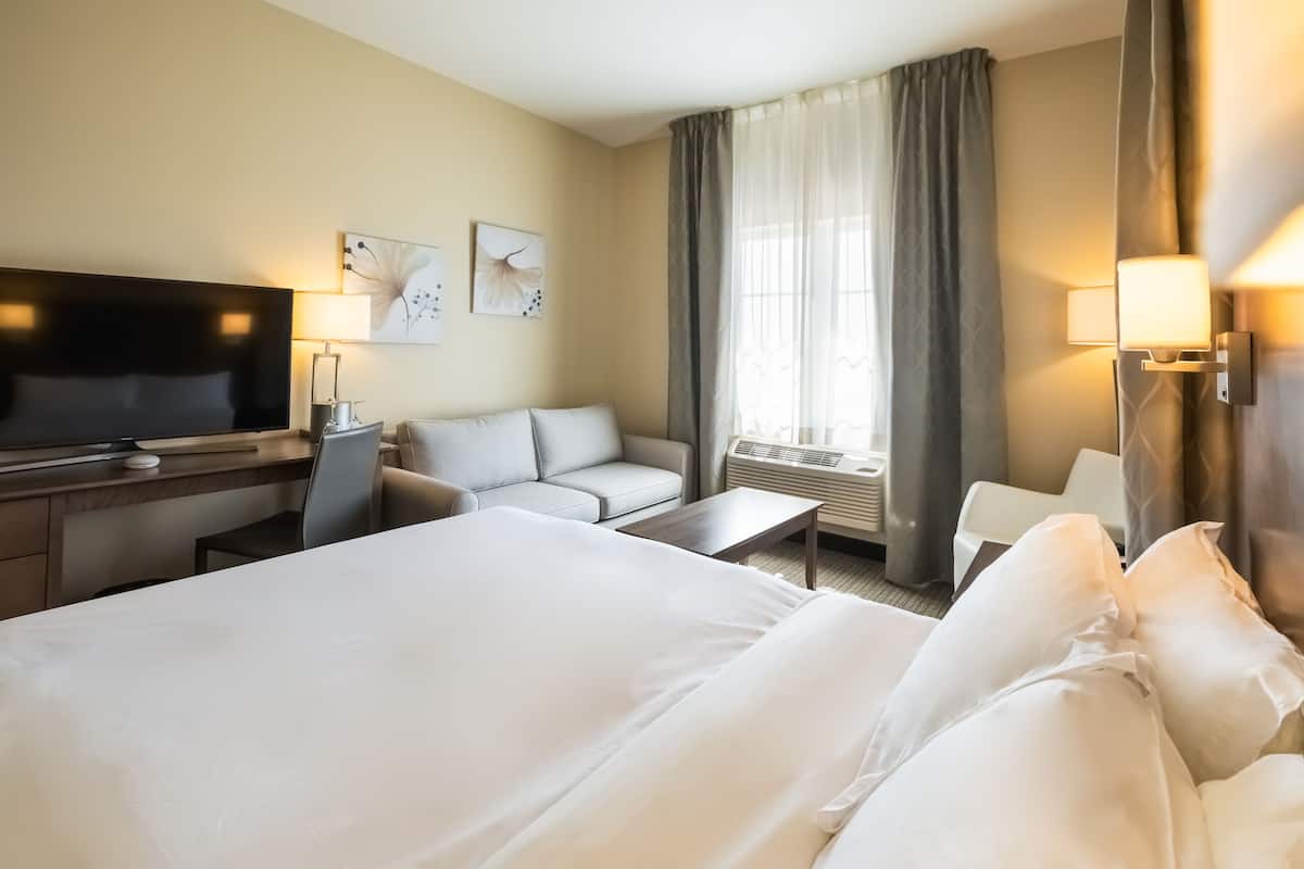 A hotel room with a large bed, a desk with a flat-screen TV, a sofa, a small table, and a window with curtains.