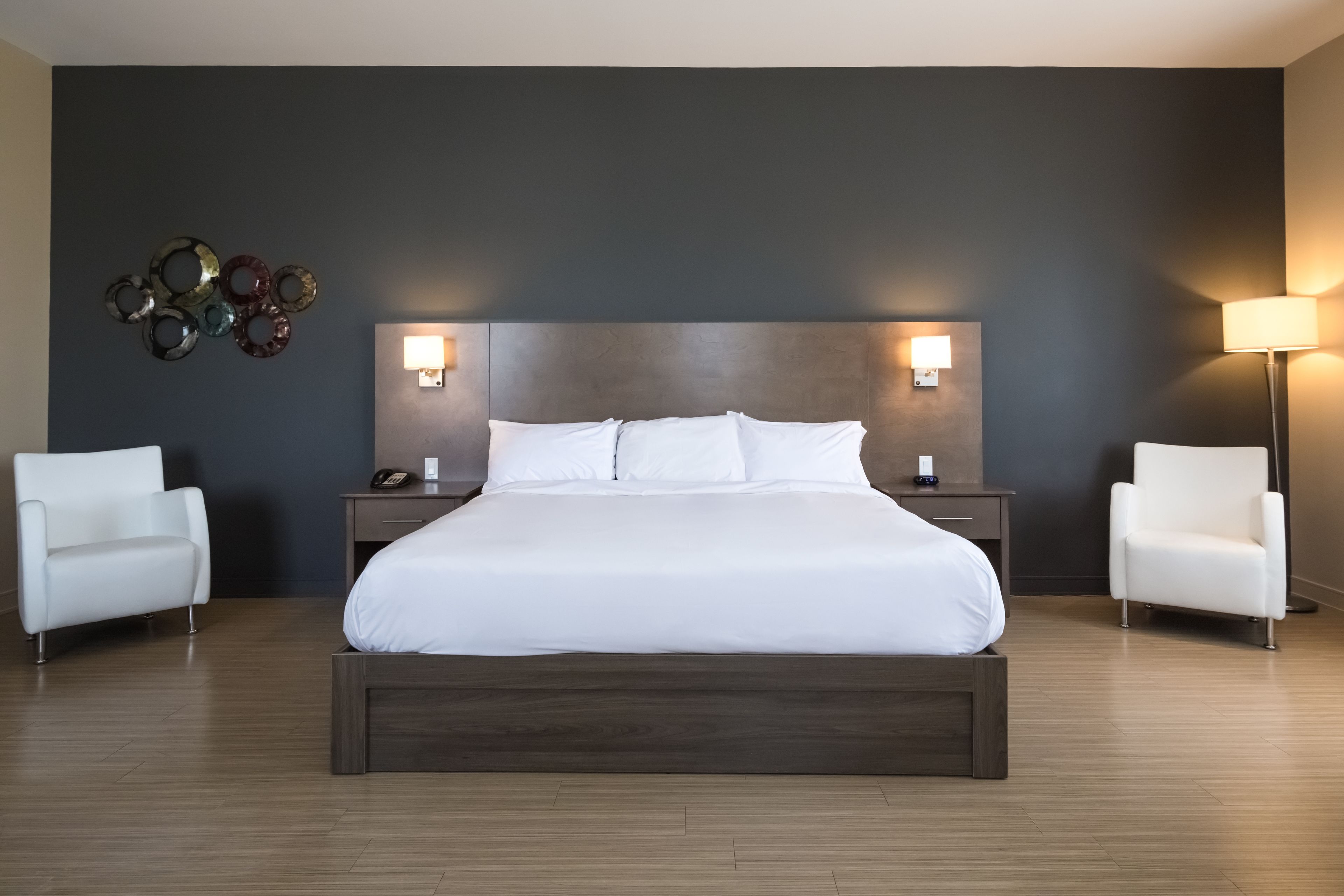A modern hotel room with a large bed, two bedside tables, a wall-mounted lamp, and two white armchairs.