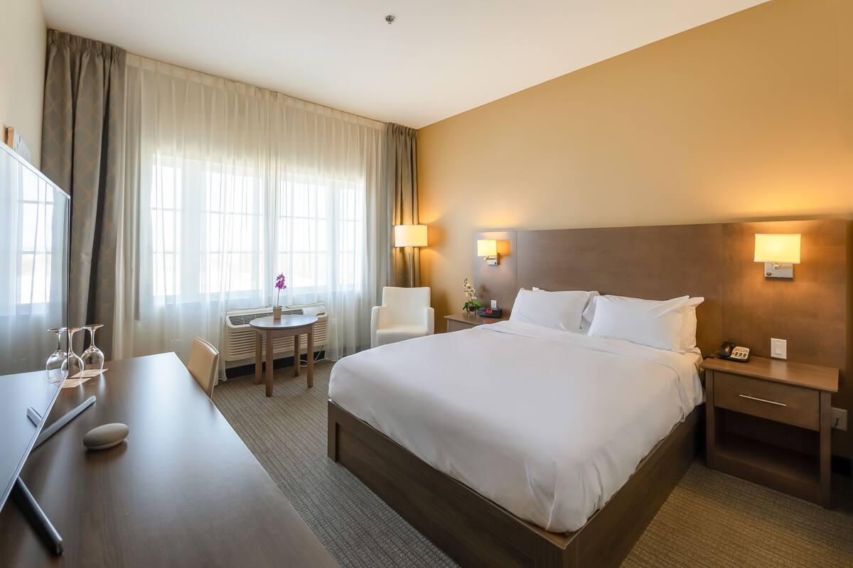A hotel room with a large bed, bedside tables, a desk with a chair, a small table with a vase, and a window with curtains.