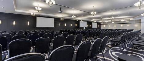 Un salón de conferencias con filas de sillas, una pantalla de proyector y varios candelabros.