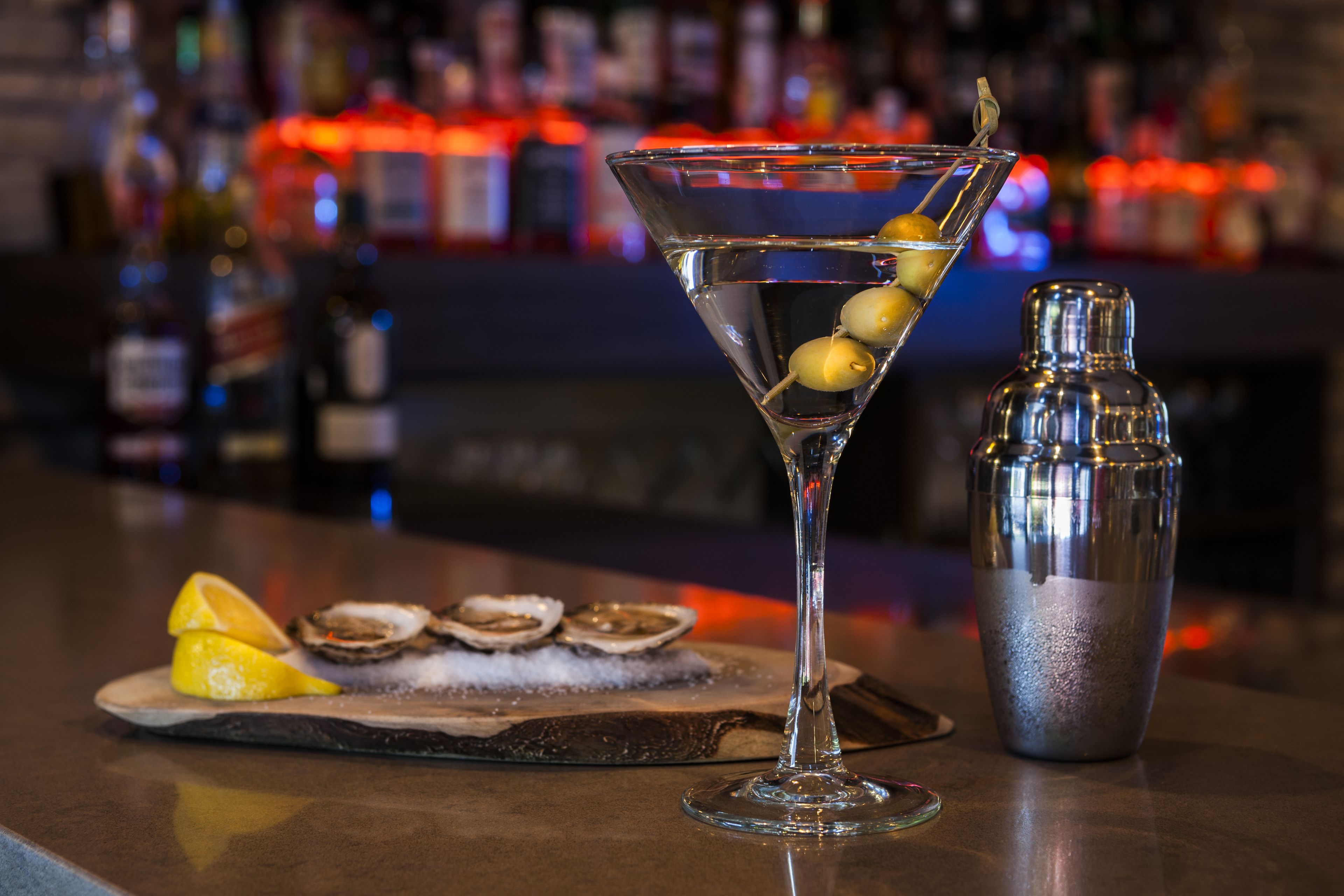 A martini glass with olives and lemon wedges on a bar counter, accompanied by a cocktail shaker.