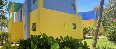 A two-story building with a yellow and blue facade, surrounded by lush greenery and palm trees.
