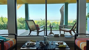 Una habitación con un ventanal grande, dos sillas, una mesa con comida y bebidas, y una cama con ropa de cama colorida.