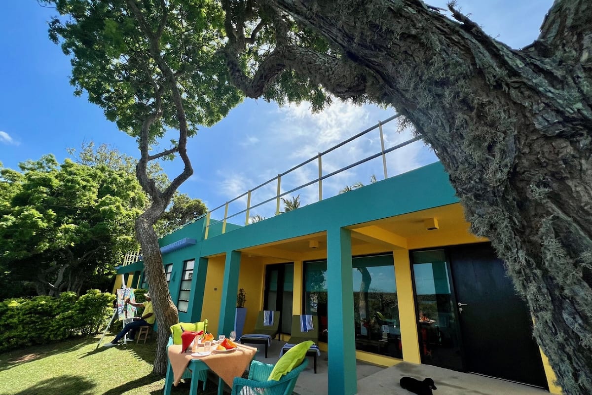 Un edificio moderno con balcón, mobiliario exterior y un árbol grande al frente.