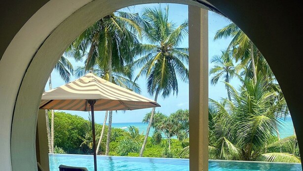 Vista circular desde un ventanal a una terraza de madera con una silla de descanso y una sombrilla, con vistas a una piscina azul cristalina y palmeras.