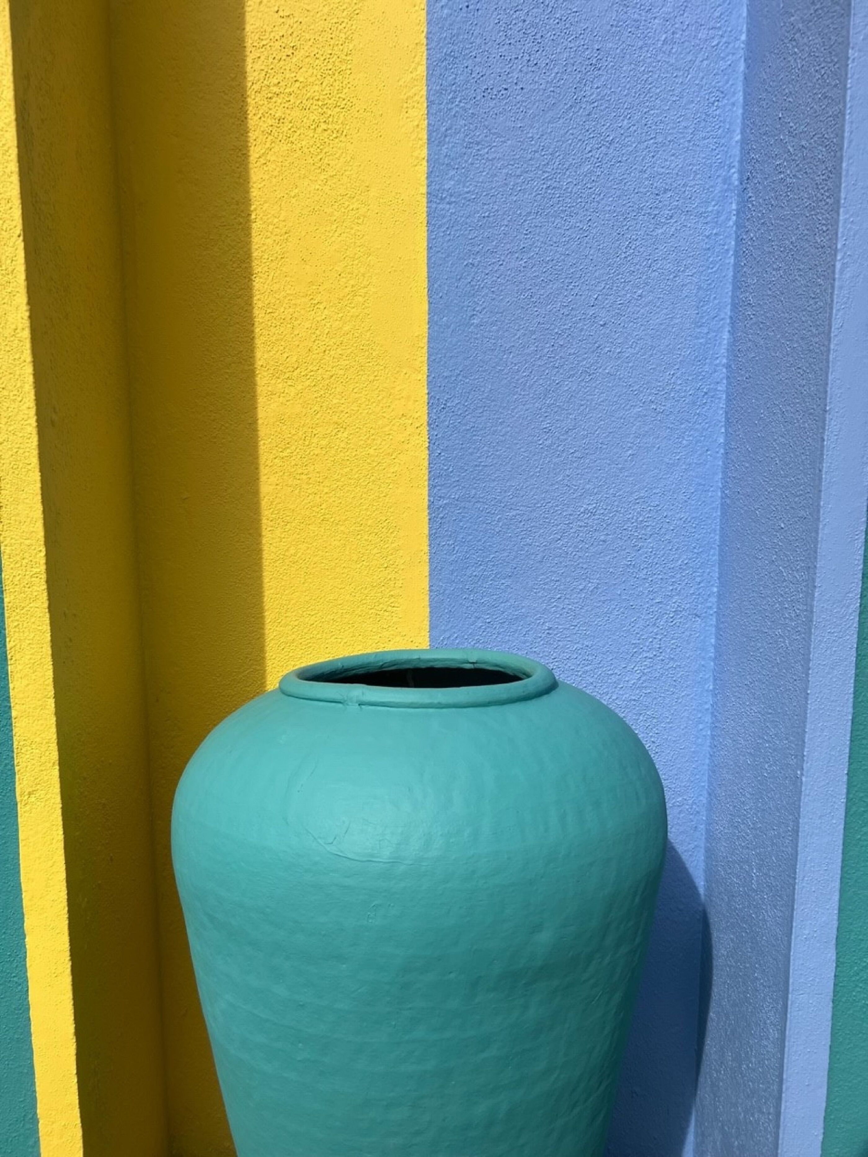 Un vaso verde grande appoggiato a un muro con strisce verticali gialle e blu.