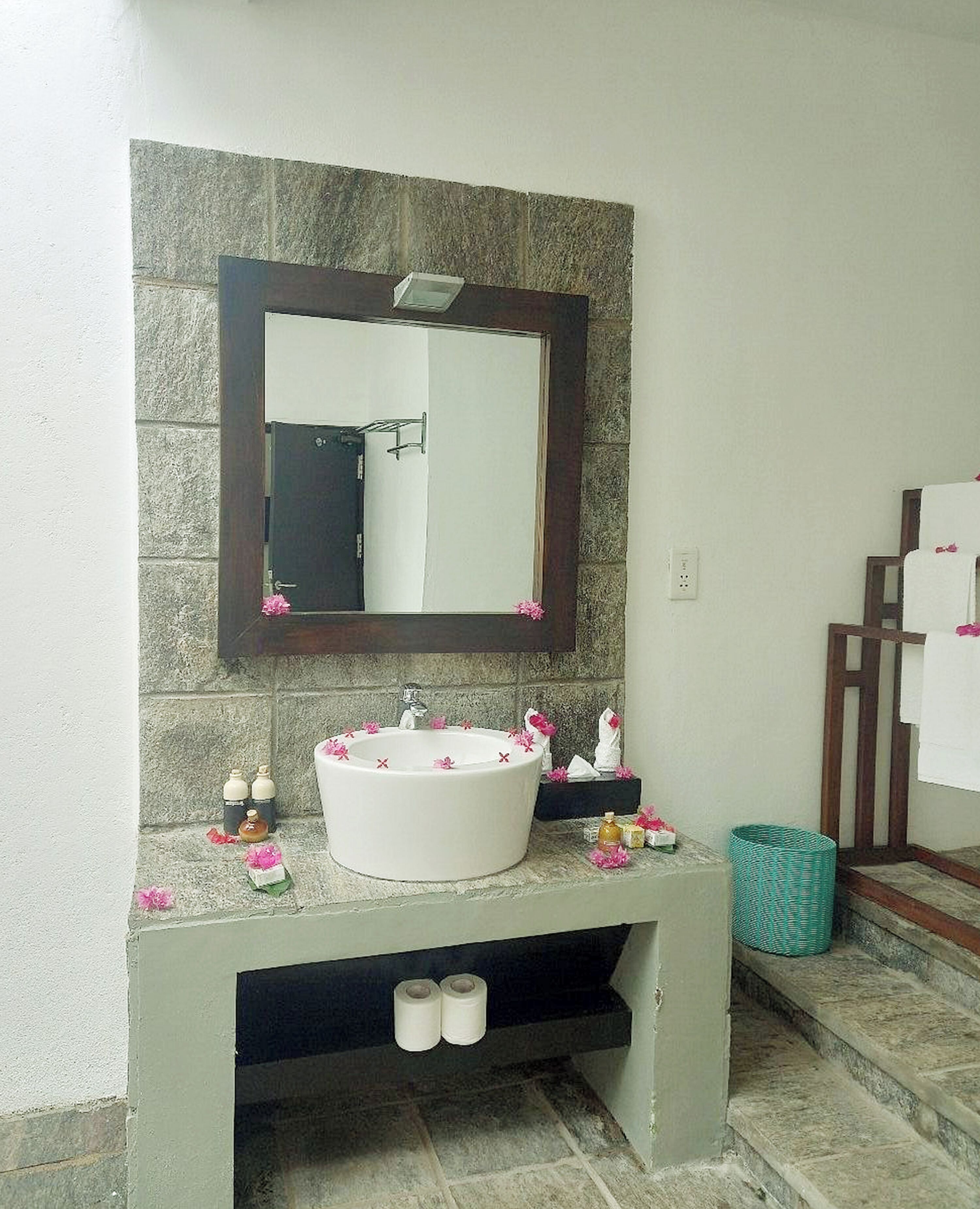 Une salle de bain avec un mur en pierre, un lavabo blanc, un miroir et une poubelle verte.