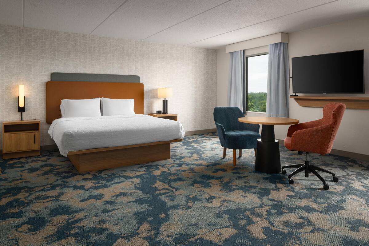 A hotel room with a bed, two chairs, a TV, and a window with curtains.