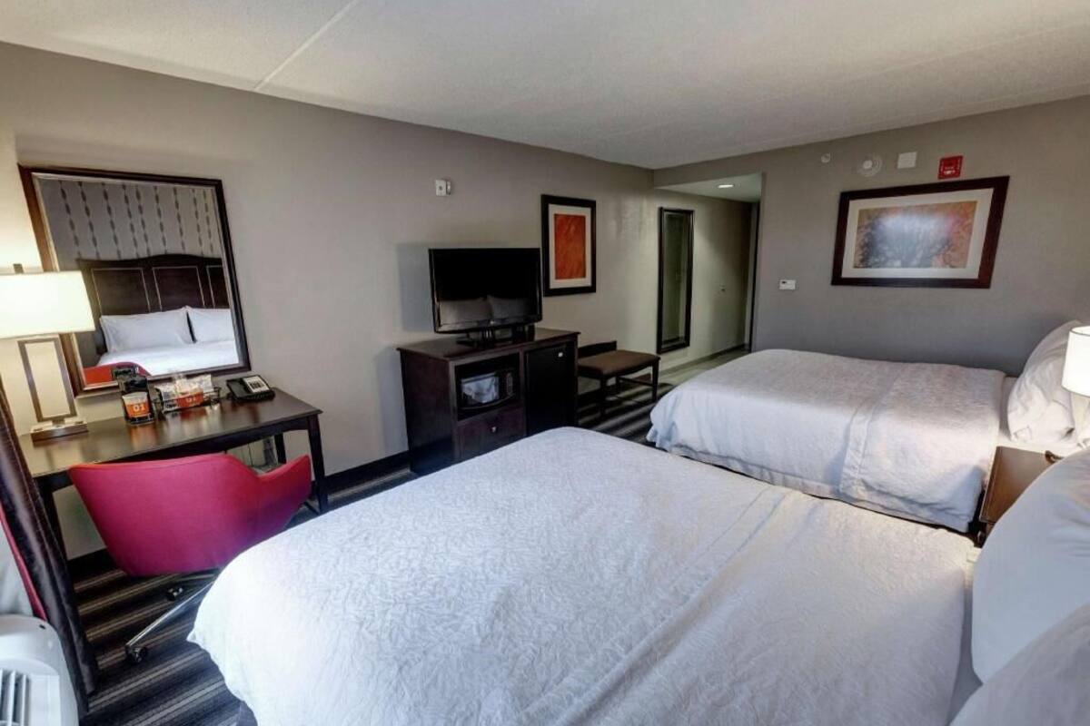 A hotel room with two beds, a desk with a TV, a chair, and a small table with amenities.