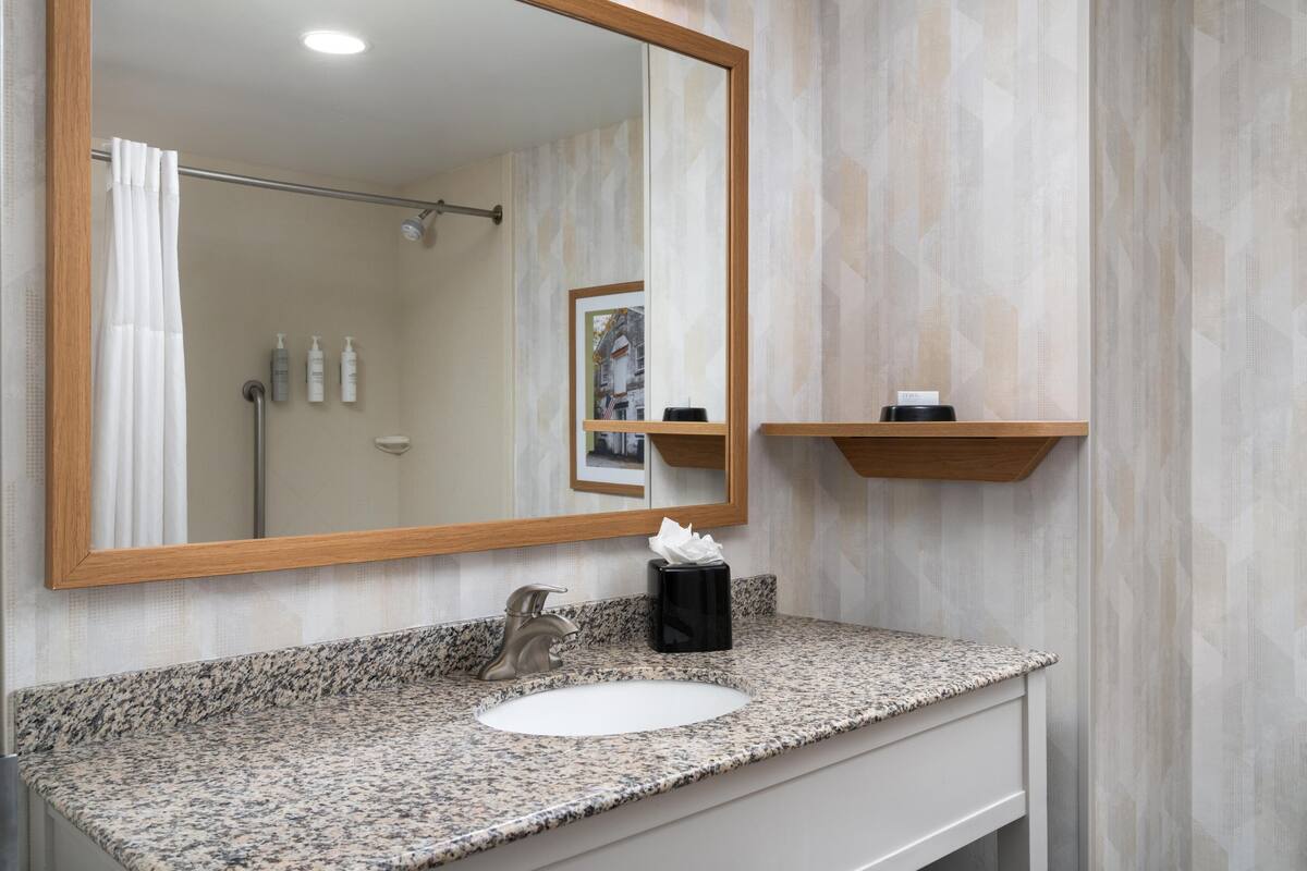 A bathroom with a granite countertop, a white sink, a mirror with a wooden frame, a towel dispenser, and a shower curtain.