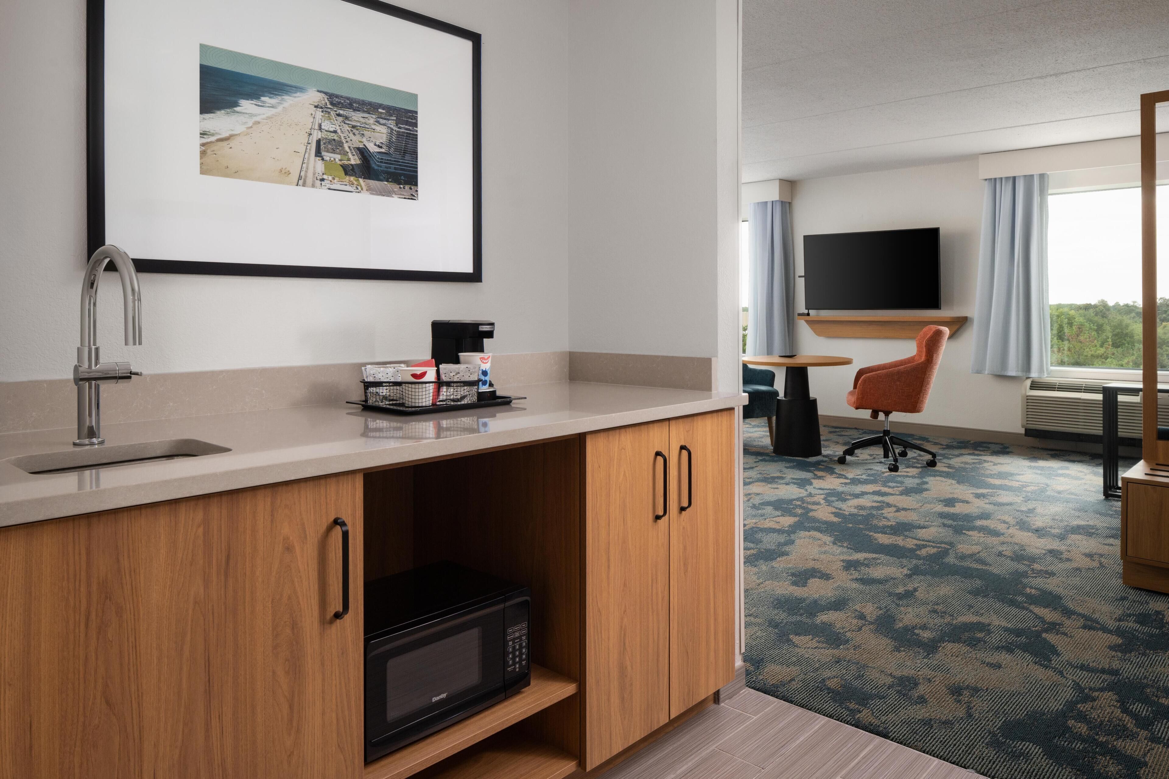 Een hotelkamer met een kitchenette, een flatscreen televisie, een raam met gordijnen en een inlijsting foto aan de muur.