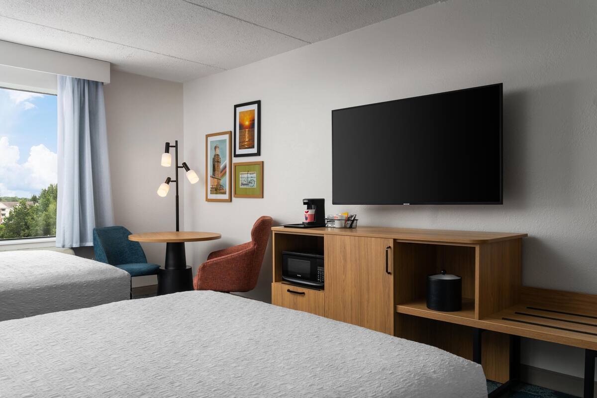 A hotel room with two beds, a desk, a chair, a TV, a microwave, and artwork on the wall.
