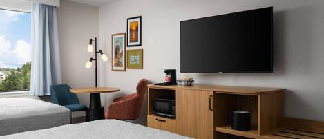 A hotel room with two beds, a desk, a chair, a TV, a microwave, and artwork on the wall.