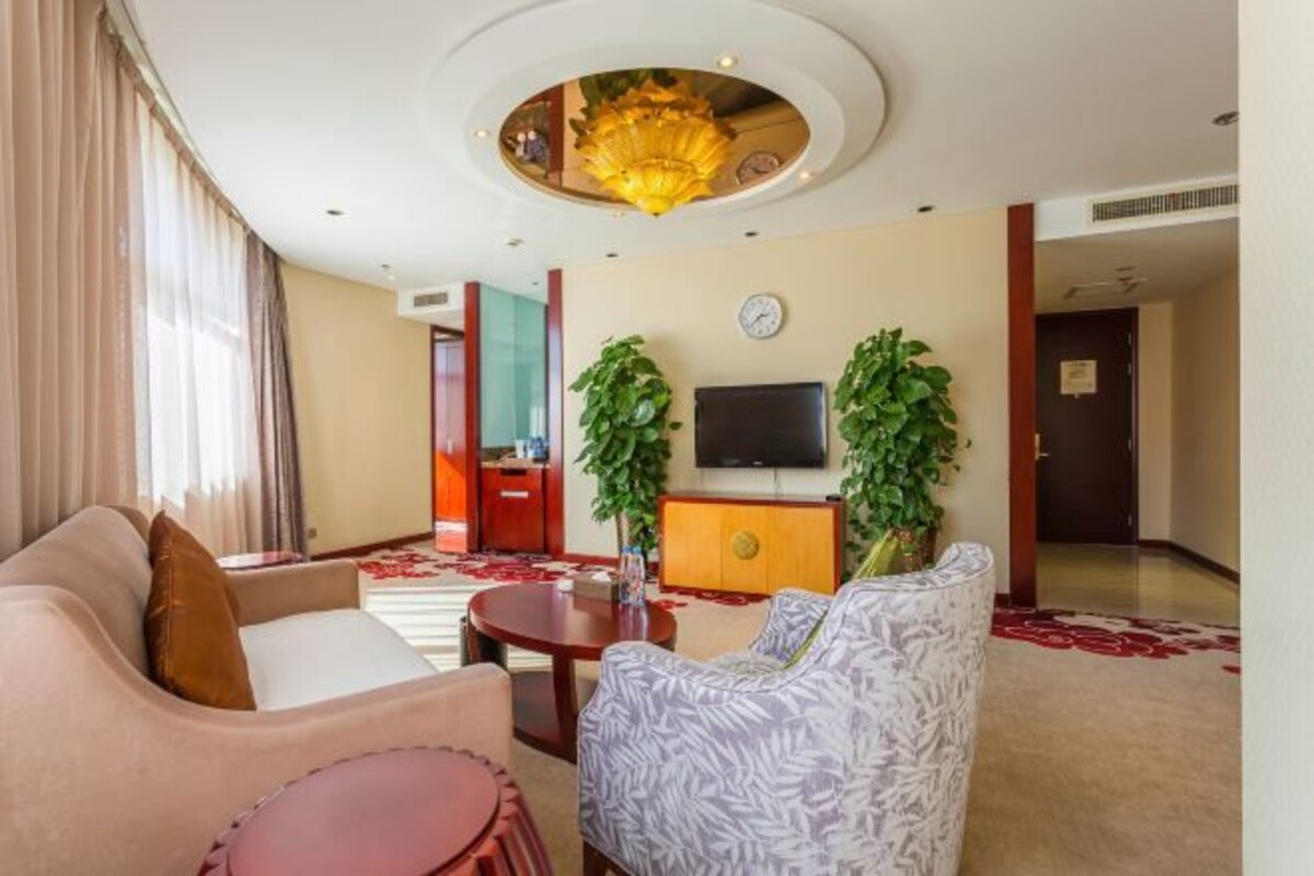 A hotel lobby with a sofa, armchair, and a small table. There is a TV mounted on the wall and two potted plants.