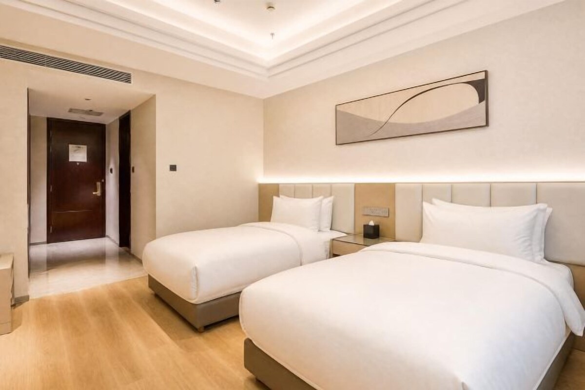 Habitación de hotel con dos camas, una mesita de noche, una lámpara de pared y una puerta que da a otra habitación.