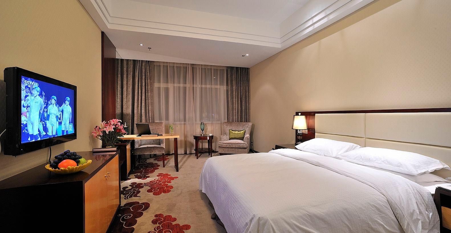 A hotel room with a bed, a desk with a chair, a television, and a window with curtains.