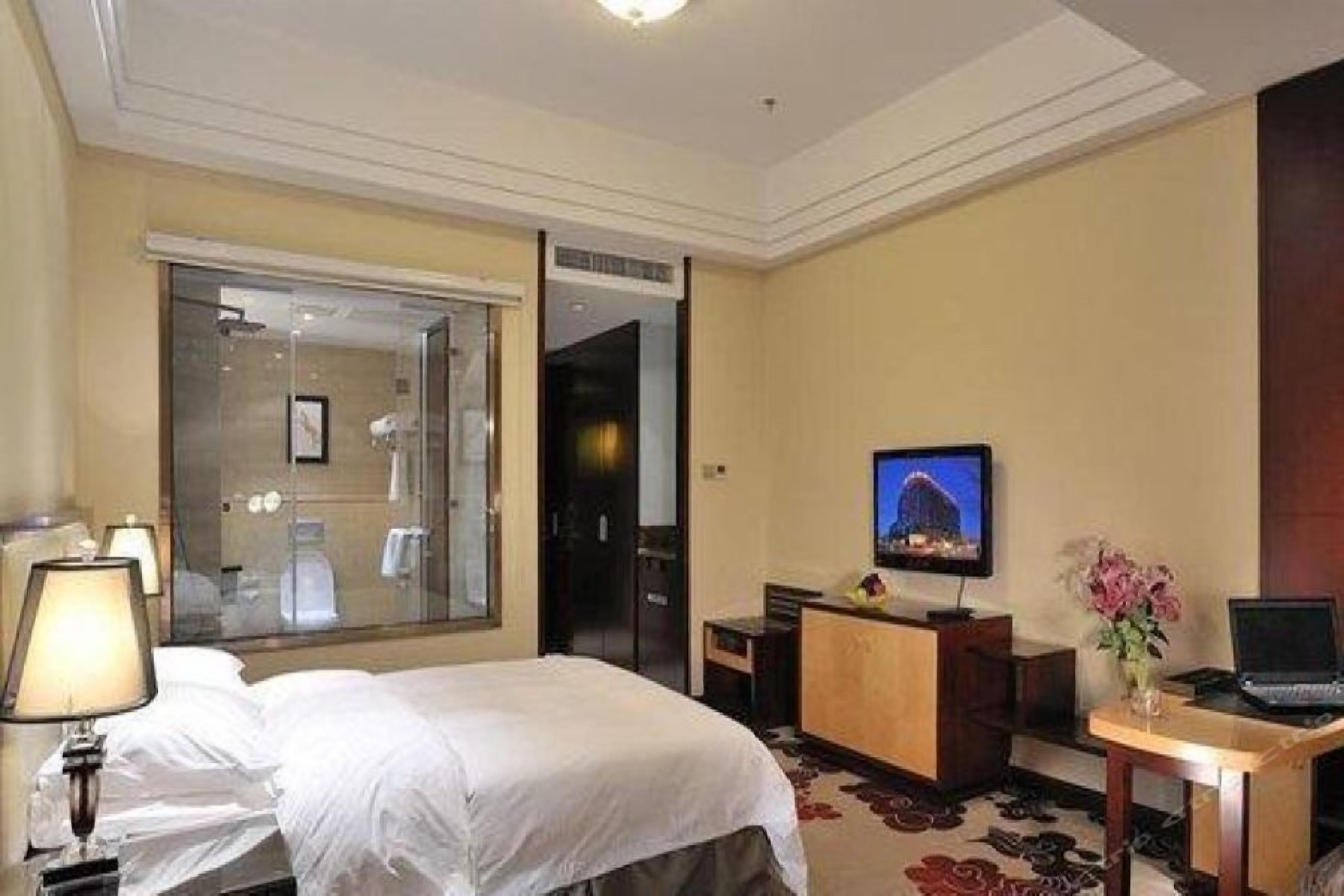 A hotel room with a bed, a desk with a laptop, a television, and a glass shower enclosure.