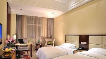 A hotel room with two beds, a desk with a lamp, a chair, and a large window with curtains.
