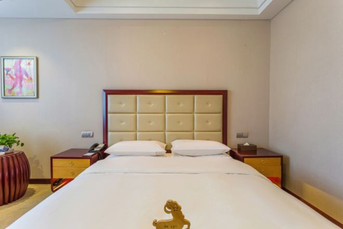 A hotel room with a large bed, a wooden headboard, two bedside tables, and a small plant.
