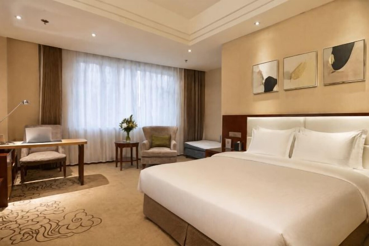 A hotel room with a large bed, a desk, a chair, and a small table with a vase of flowers.