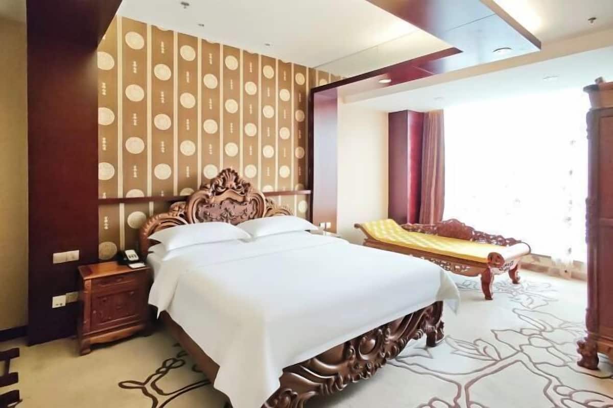 Une chambre d’hôtel avec un grand lit, une table de chevet, une tête de lit en bois, un papier peint à motifs et une fenêtre avec des rideaux.
