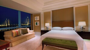 A hotel room with a large bed, a green upholstered bench, a sofa with colorful pillows, and a night view of a bridge and city lights. - Sheraton Fuzhou Hotel (Fuzhou)