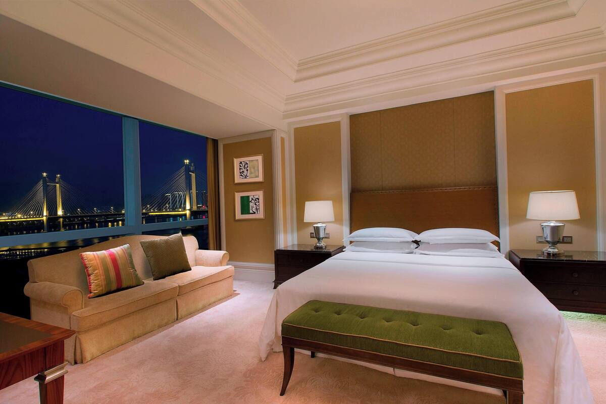 A hotel room with a large bed, a green upholstered bench, a sofa with colorful pillows, and a night view of a bridge and city lights.