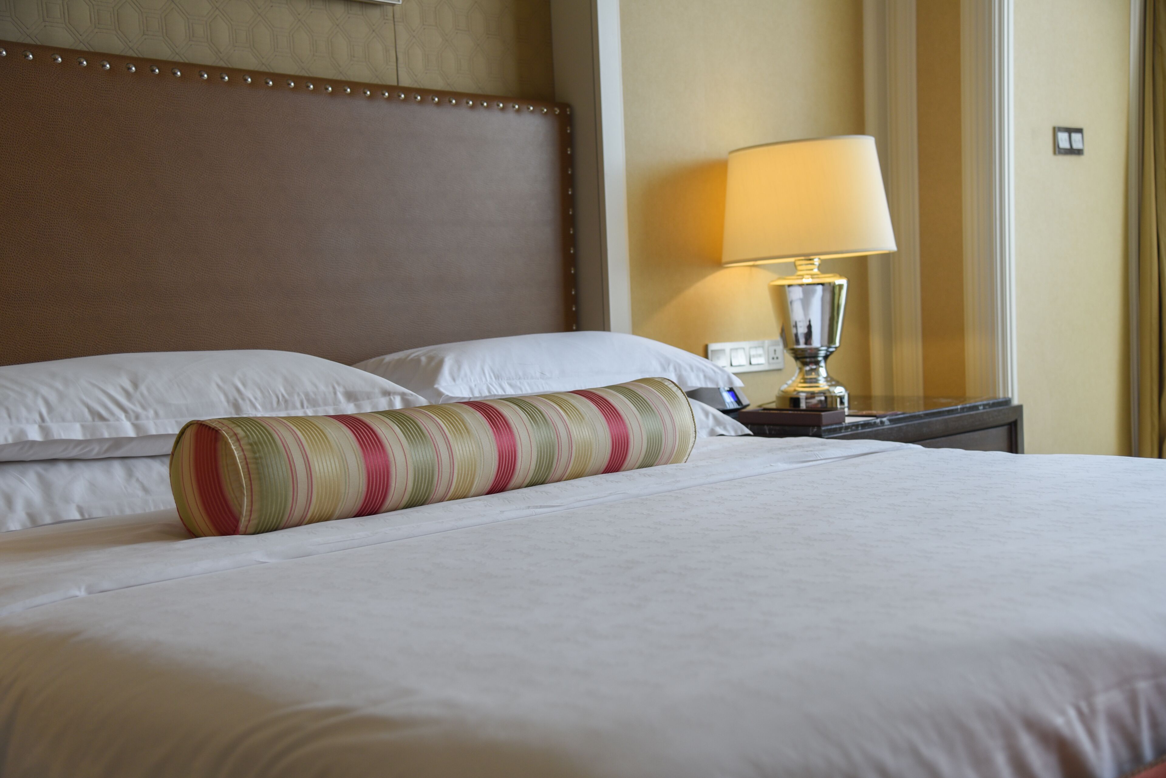 Una cama bien hecha con sábanas blancas y un cojín a rayas, una lámpara de noche y una pared con un diseño decorativo.