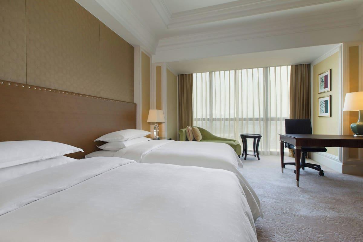 A hotel room with two large beds, a desk, a chair, and a large window with curtains.