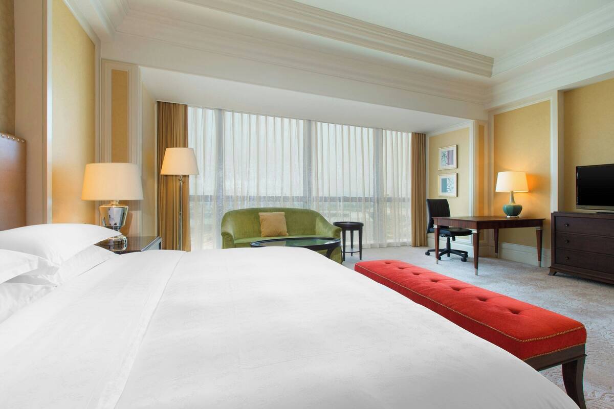 A hotel room with a large bed, a green armchair, a red ottoman, a desk with a lamp, a television, and a view of the city through large windows.