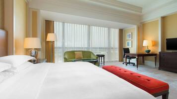 Une chambre d’hôtel avec un grand lit, un fauteuil vert, un pouf rouge, un bureau avec une lampe, une télévision et une vue sur la ville grâce à de grandes fenêtres.