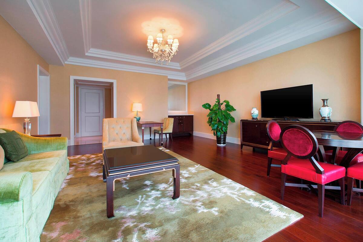 A spacious living room with a large television, a chandelier, and a dining area with red chairs.