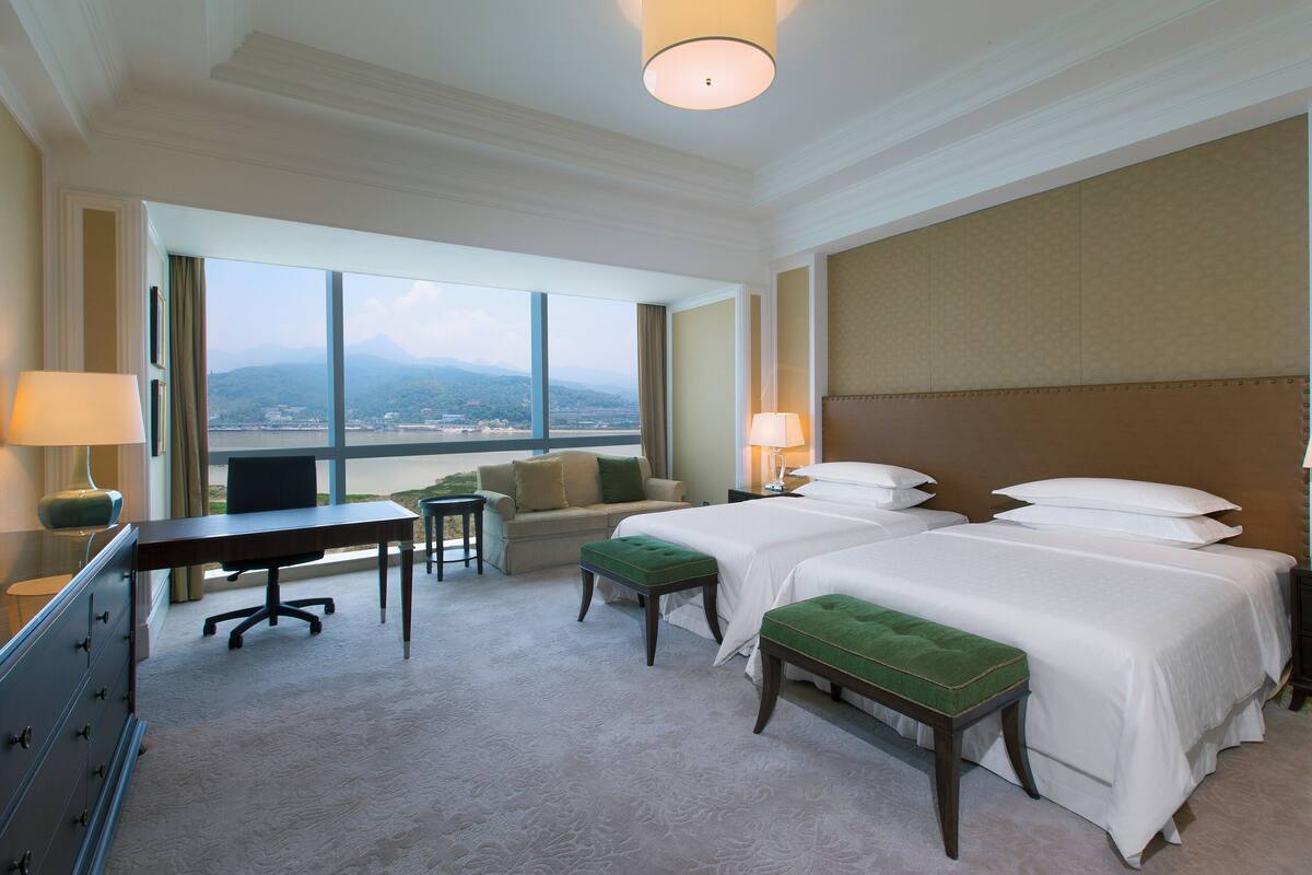 A hotel room with two beds, a desk, a chair, a sofa, and a view of the mountains.