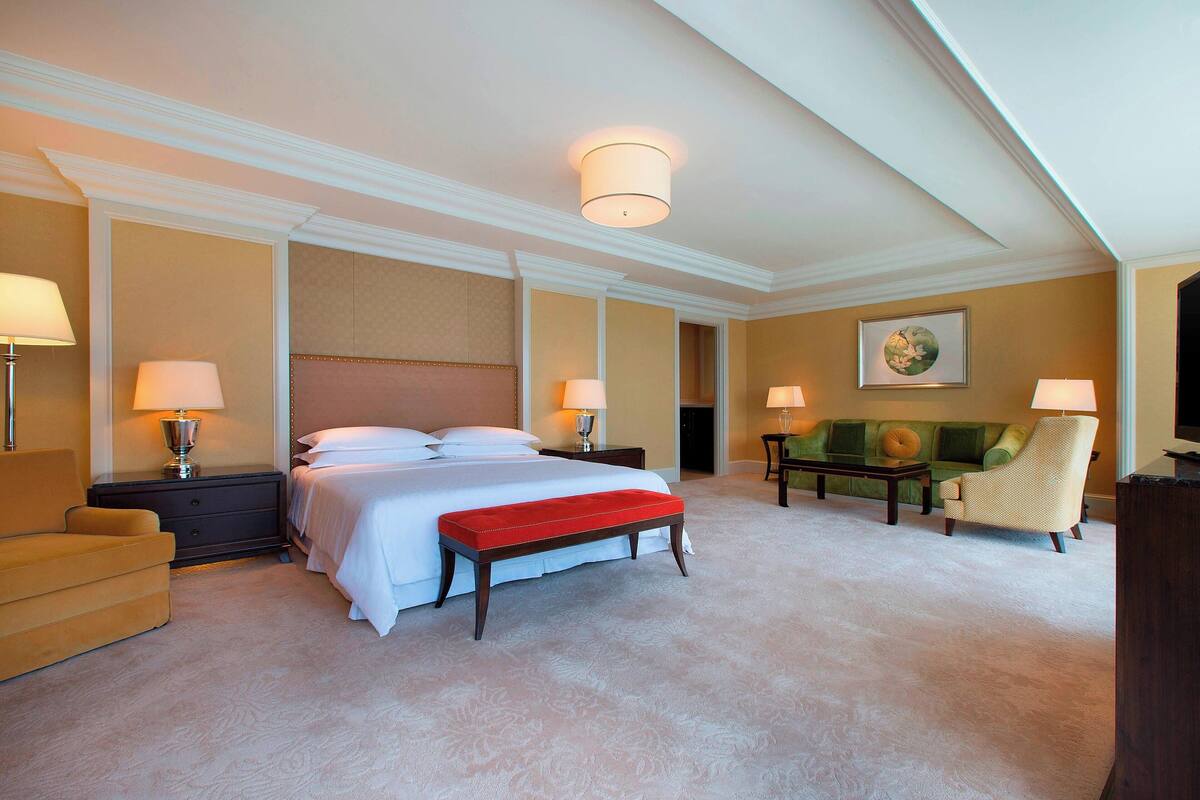 A spacious hotel room with a large bed, a red bench, a green sofa, and a wooden nightstand with lamps.