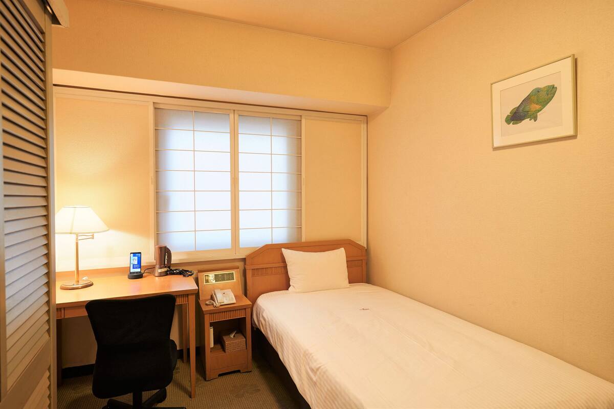 Quarto de hotel com uma cama de solteiro, uma secretária com cadeira, uma janela com persianas, uma mesa de cabeceira com abajur e um quadro pendurado na parede.