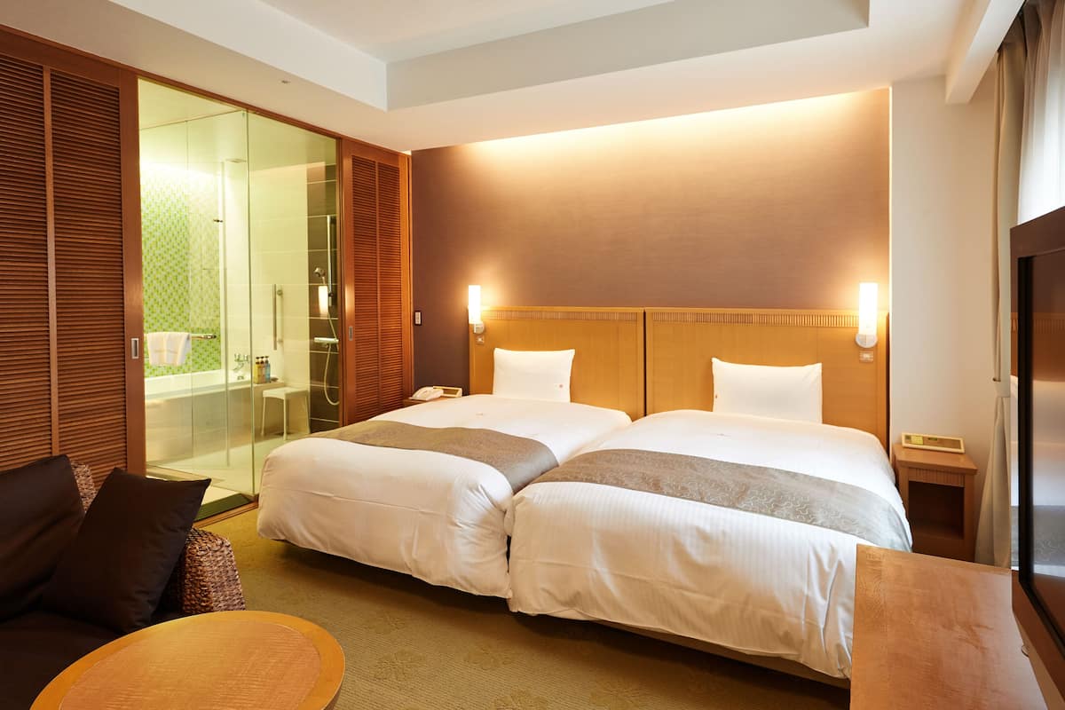 Quarto de hotel com duas camas, um sofá, uma mesa pequena e área de chuveiro com divisória de vidro.
