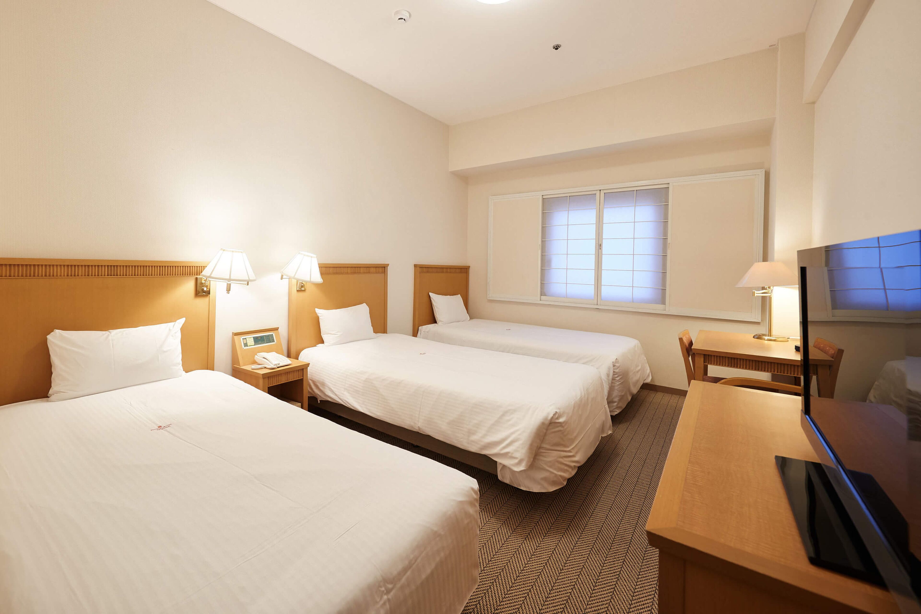 A hotel room with three single beds, a wooden desk, a television, and a window with blinds.
