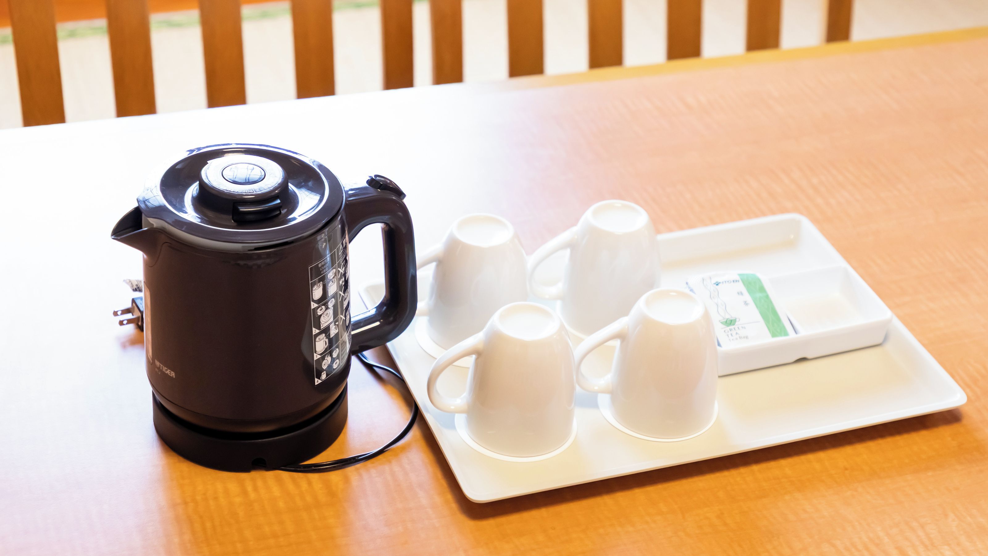 Une bouilloire électrique noire est posée sur une table, accompagnée d’un plateau blanc contenant quatre tasses blanches et d’une boîte de thé vert.