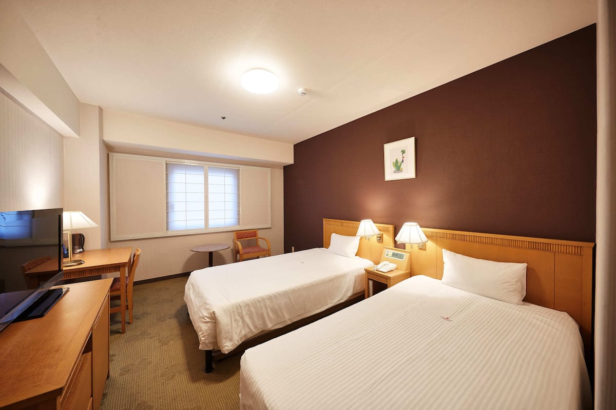 Quarto de hotel com duas camas, uma escrivaninha, uma cadeira, uma televisão e uma janela com persianas.